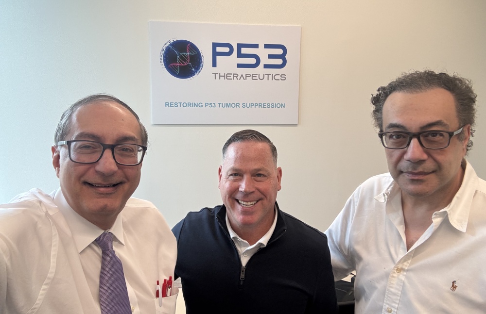 Three men posing for a photo in an office with a P53 Therapeutics sign in the background; central figure smiles wearing a black zip sweater, flanked by two men in white shirts.