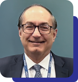 Smiling man in a dark suit and tie, wearing glasses and a blue lanyard, against a teal background.